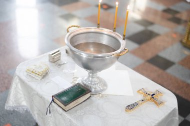 Baptismal font with blessing water, wax candles and cross are on the table covering by white tablecloth near Holy Bible. Newborns baptism rite in Orthodox church.