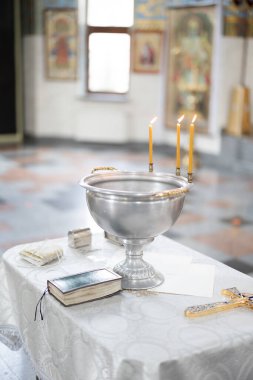 Baptismal font with blessing water, wax candles and cross are on the table covering by white tablecloth near Holy Bible against icons background. Newborns baptism rite in Orthodox church.