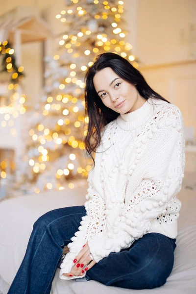 Young pretty woman in white sweater and jeans sits on bed against background of Christmas tree and glowing garlands with bokeh effect.