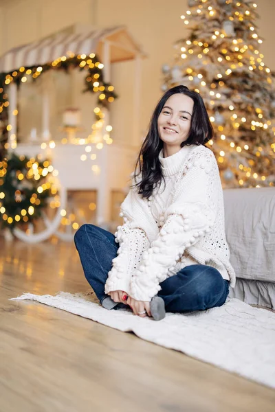 Young cheerful woman in white sweater and jeans sits on floor against background of Christmas tree and glowing garlands.