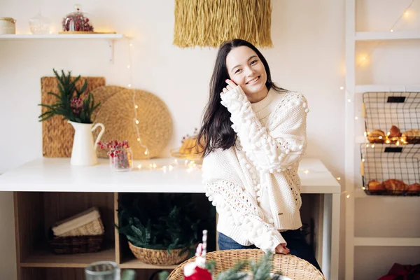 Young joyful woman in white sweater and jeans stands in kitchen against background of Christmas glowing garlands and decorations.