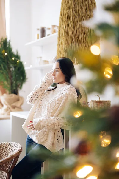 Young pensive woman in white sweater and jeans stands in kitchen against background of Christmas tree and glowing garlands.