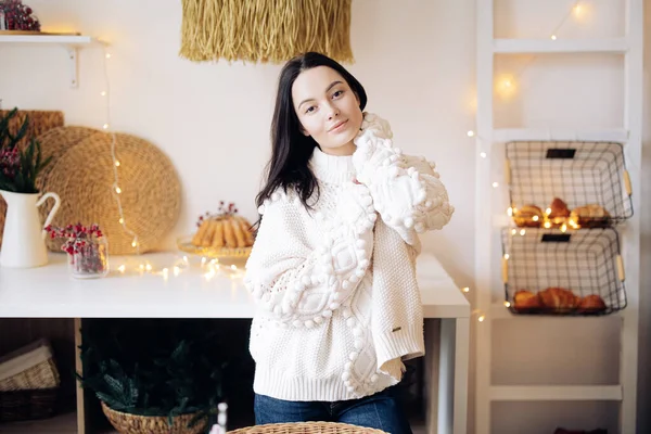 Young pensive woman in white sweater and jeans stands in kitchen against background of Christmas glowing garlands and decorations.