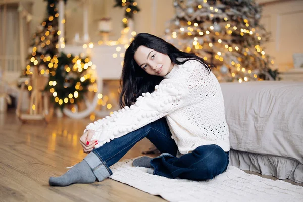 Young smiling woman in white sweater and jeans sits on floor against background of Christmas tree and glowing garlands with bokeh effect.