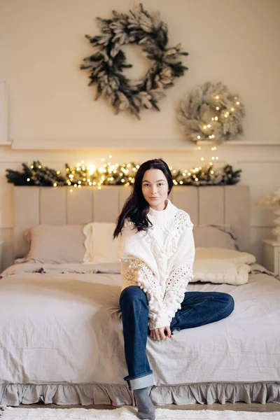 Young pretty woman in white sweater and jeans sits on bed against background of Christmas wreath and glowing garlands.