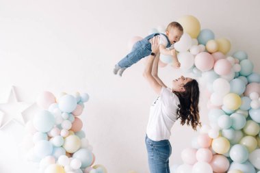 Happy mother has fun and plays with her son celebrating first birthday of baby boy among a lot of colorful balloons. Celebration first birthday. Festive event.