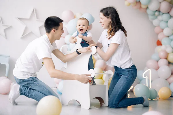 Happy parents have fun and play with their son celebrating first birthday of baby boy among a lot of colorful balloons. Celebration first birthday. Festive event.