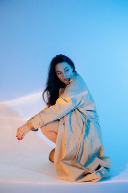 Genç, güzel esmer kadın mavi arka planda düğmesiz pelerin giymiş poz veriyor..