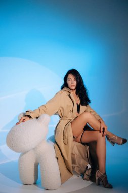 Genç seksi kadın siyah dantelli iç çamaşırlarıyla BDSM tarzında oturuyor ve mavi arka planda düğmesiz pelerin giyiyor..