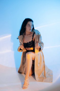 Genç seksi kadın siyah dantelli iç çamaşırlarıyla, yüksek topuklu ayakkabılarıyla ve mavi arka planda düğmesiz peleriniyle oturuyor..