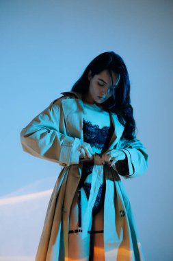 Genç seksi kadın siyah dantelli iç çamaşırlarıyla BDSM tarzında poz veriyor ve mavi arka planda düğmesiz pelerin giyiyor..