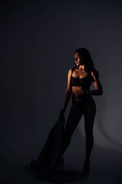 Genç seksi kadın elinde pelerinle siyah dantelli iç çamaşırlarıyla BDSM tarzında poz veriyor. Aydınlandı..