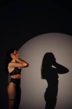 Genç seksi kadın siyah dantelli iç çamaşırlarıyla BDSM tarzıyla duvardaki gölgesinin arkasına poz veriyor. Aydınlandı..