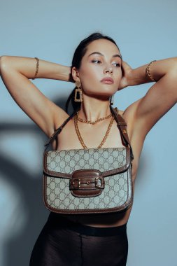 Genç ve güzel bir kadın üstsüz poz veriyor ve gri arka planda el çantasıyla üstünü örtüyor..