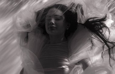 Güneş ışınlarında suda dinlenen şeffaf elbiseli güzel bir kadının sualtı çekimi. Fantezi deniz kızı, arka planda su yüzeyine karşı ışık huzmeleriyle. Siyah beyaz görüntü. Yakın çekim..