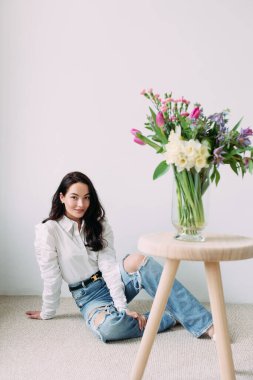 Genç ve mutlu bir kadın, duvarın arkasında, masanın üzerinde güzel bir buket çiçeğin yanında oturuyor. Anneler günü, doğum günü, yıldönümü, kutlama, tatil, selamlama, bayram etkinliği.