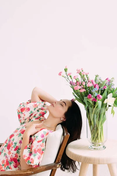 テーブルの上に美しい花の花束と花瓶の横に閉じ目でアームチェアに座って若い夢の女性 母の日 誕生日 記念日 お祝い お祝いイベント — ストック写真