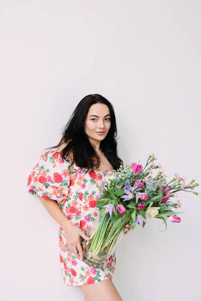 stock image Young happy woman standing and holding vase with flowers bouquet against white background. Mothers day, birthday, anniversary, celebration, holiday, greeting, festive event. Isolated image.