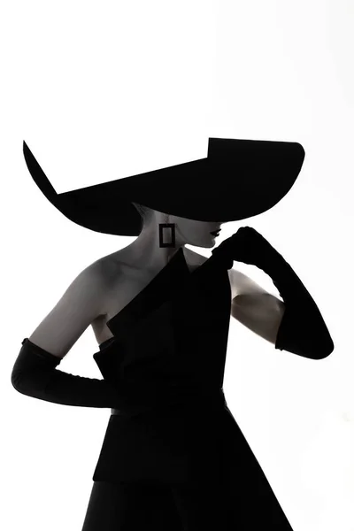 白い背景を背景に スタイリッシュでつばの広い帽子 黒いドレス 手袋をしたスタジオで化粧をした若い女性がポーズをとっている 孤立したイメージ サイドビュー — ストック写真