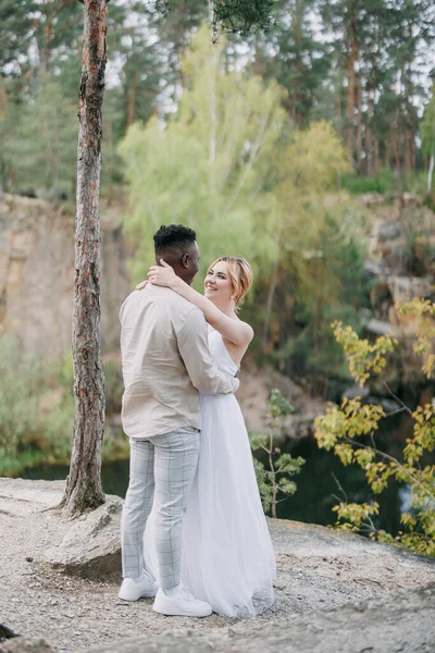 stock image Happy interracial couple newlyweds stands, embraces and smiles against background of lake and forest. Concept of love relationships and unity between different human races.