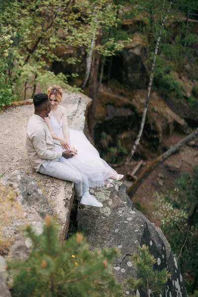 若い異人種間のカップル新婚夫婦は岩の上に座って 森林や峡谷の背景を背景に話をします 異なる人種間の愛の関係と団結の概念 — ストック写真