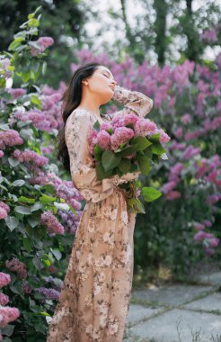 Genç, güzel esmer kadın elinde buketle bahçede yürüyor ve çiçek açan leylak çalılarıyla gözleri kapalı eğleniyor..