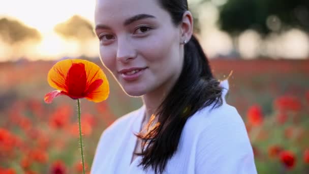 彼女の手の中にケシの花を持つ若い女性の肖像画笑顔と日没時に牧草地を開花によって楽しむ — ストック動画