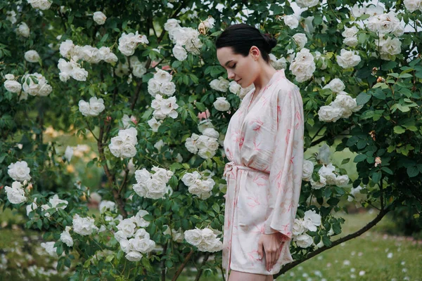 着物姿の若いブルネットの女性が白アイスバーグを開花させることで楽しむ庭でバラ — ストック写真
