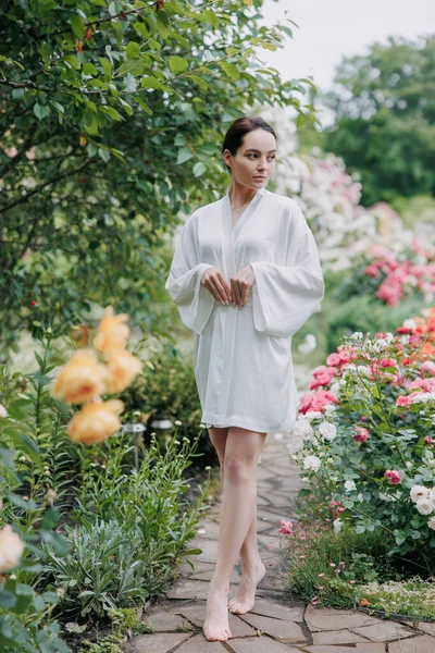 stock image Young brunette woman in kimono dress walking barefoot on flagstone paved path among roses in garden and enjoying by bloom.