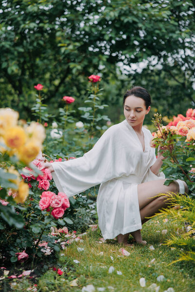 Young brunette woman in kimono dress sitting barefoot on lawn among roses in garden and enjoying by bloom.