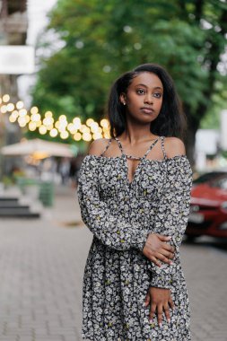 Yazlık elbiseli genç Afrikalı düşünceli kadın kentte dikiliyor ve sokak arkasında poz veriyor..