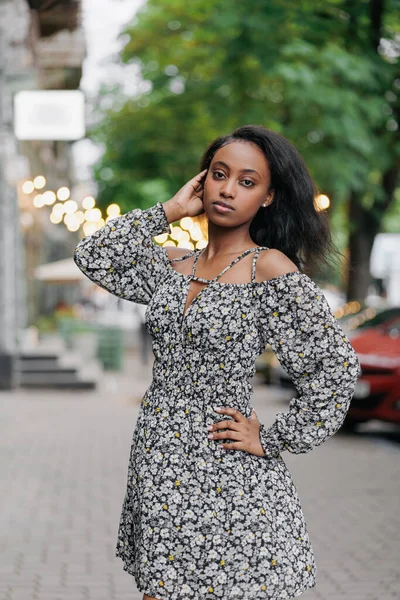 stock image Young african pensive woman in summer dress stands and poses in city against background of street.