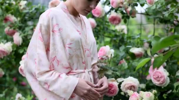 Młoda Kobieta Wiąże Pasek Peignir Ogrodzie Wśród Kwitnącej Różowej Róży — Wideo stockowe