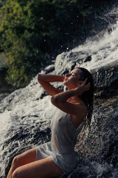 濡れたドレスを着た若い女性が岩の上に座って 水の流れ スプレー 泡の間の滝の近くで閉じた目で楽しんでいます — ストック写真