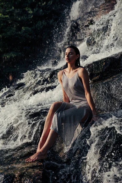 岩の上に座って 水の流れ スプレーや泡の間の滝の近くに閉じた目で楽しむぬれたドレスの若い女性 — ストック写真