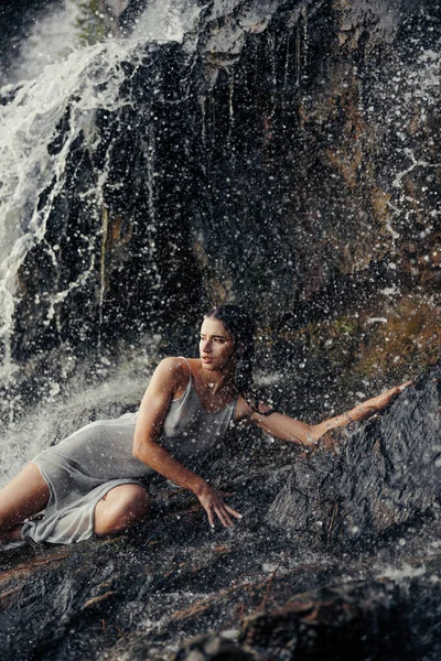 岩の上に横たわる濡れたドレスの若い女性と水の流れ スプレーや泡の間の滝の近くを楽しんで — ストック写真