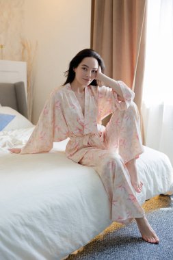 Pijamalı genç esmer kadın yatak örtüsü ve pencerenin arkasında oturuyor..