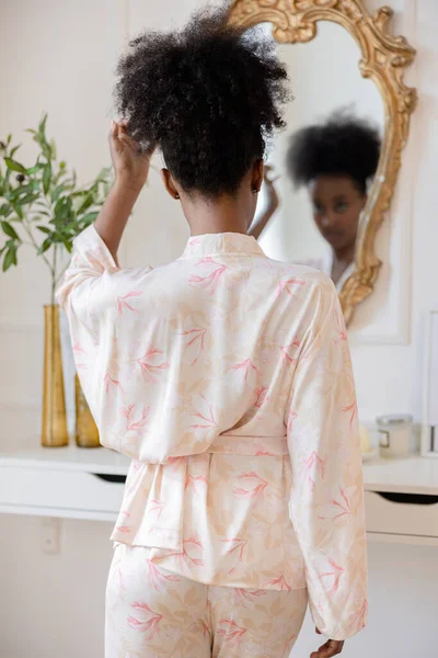 鏡の前に立つパジャマ姿の若いアフリカ人女性は 家の中を背景にその姿を見つめます バックビュー — ストック写真