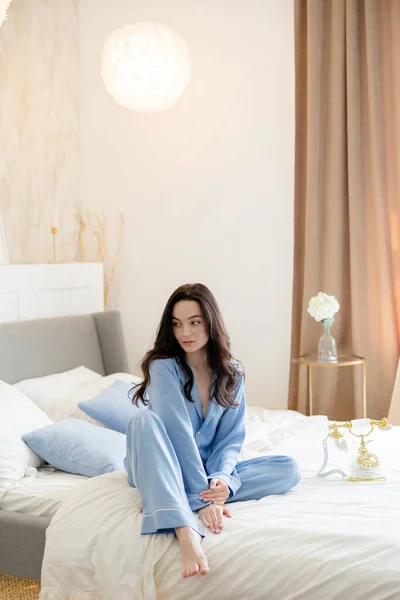 若いですPensiveブルネット女性で青パジャマでベッドの上に座っている背景にベッド服と家のインテリア — ストック写真