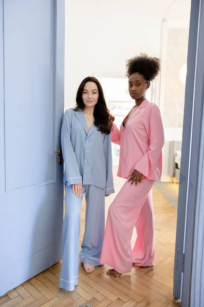 stock image Couple of african and caucasian women in pajamas standing in doorway of bedroom against background of home interior.