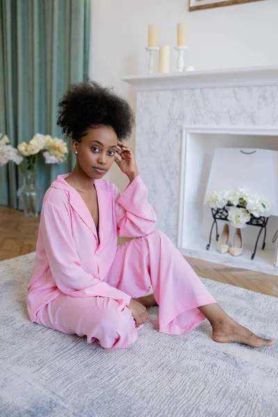 Young african woman in pink pajamas sitting on carpet in bedroom against background of home interior.