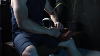 Erkek boksör, karanlık spor salonunda yedek kulübesinde oturuyor ve boks eldiveni giyiyor. Yakın çekim..