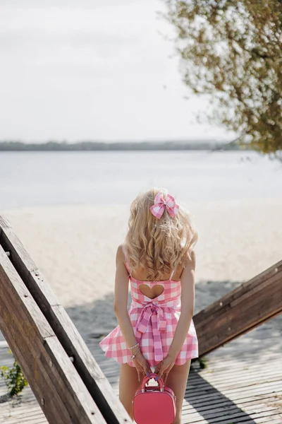 Young blonde girl look like a Barbie doll in pink checkered mini dress and pink bow in her hair with handbag standing on beach against sand and water background. Back view.