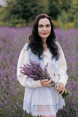Genç esmer kadın elinde bir demet lavanta ile çiçek açan lavanta tarlasının arka planında yürüyor. Yakın plan..
