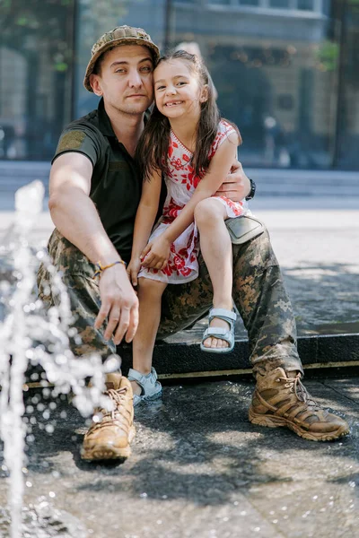 Ukrainian soldier returning from the war meets his happy little daughter and embraces her near fountain in city. War in Ukraine. Russian military invasion in Ukraine. War and children. Close-up.