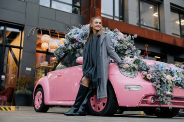 Şehrin arka planında çiçek çelenkleriyle süslenmiş pembe arabanın yanında duran mutlu genç kadın..