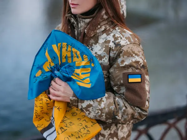Ukrainian woman soldier holding flag with signatures of Ukrainian warriors and resting in park in vacation. Translation from Ukrainian: Glory to Ukraine. Closeup.