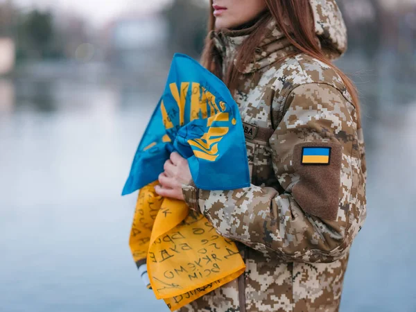 Ukrainian woman soldier holding flag with signatures of Ukrainian warriors and resting in park in vacation. Translation from Ukrainian: Glory to Ukraine, surname. Closeup.