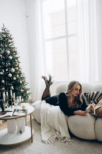 Young pensive blonde woman lying down on sofa with Christmas tree ball in her hands in stylish black dress and high heel shoes near decorated Christmas tree, table with candlestick and window.