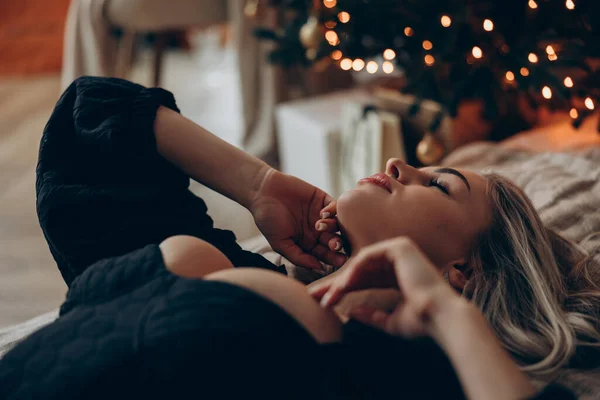 Young dreamy blonde woman lying down on bed in black decollete dress near decorated Christmas tree. Close-up.